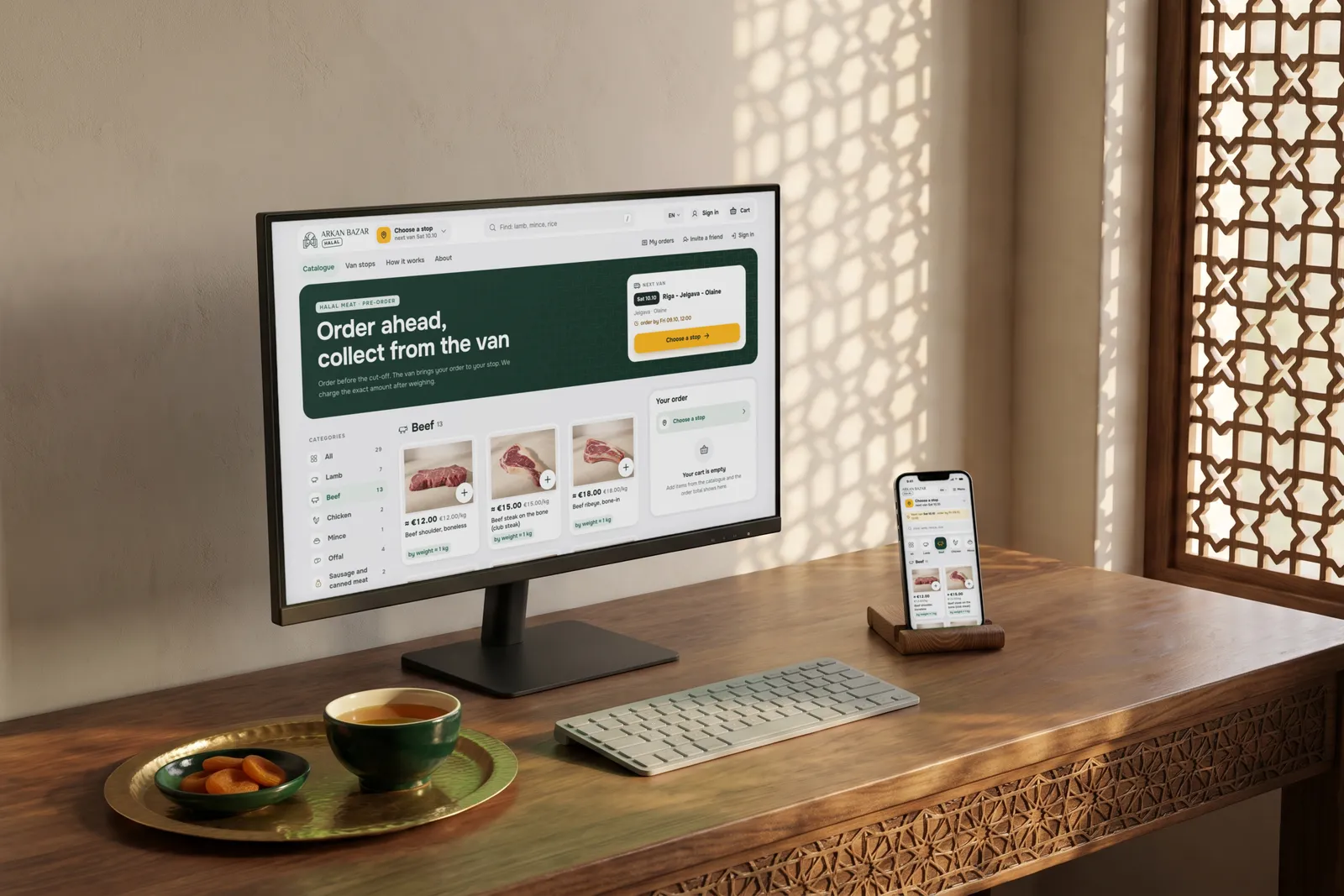 A desktop monitor and a phone on a walnut desk by a carved wooden lattice window, both showing the Arkan Bazar website in English with its beef catalogue and the "Order ahead, collect from the van" banner